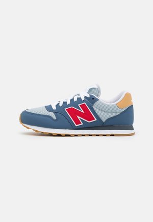Blauwe en grijze sportschoen met witte veters, rood "N" logo aan de zijkant, beige hielaccent en zool in kauwgumkleur.