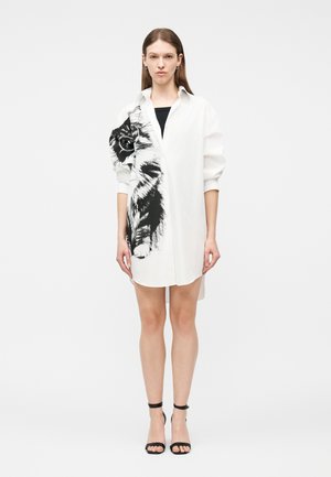 Woman stands wearing a white oversized shirt dress with a large black and white cat print on the left side, paired with black ankle-strap heels.