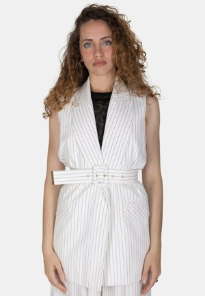 Donna con capelli ricci che indossa un gilet bianco a righe sottili senza maniche con cintura sopra una maglia nera, in piedi davanti a uno sfondo semplice.