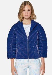 Marineblaue Steppjacke mit gl änzender Oberfläche, gestepptem Design, Spitzkragen und Reißverschluss vorne. Kombiniert mit hellblauen gemusterten Hosen.