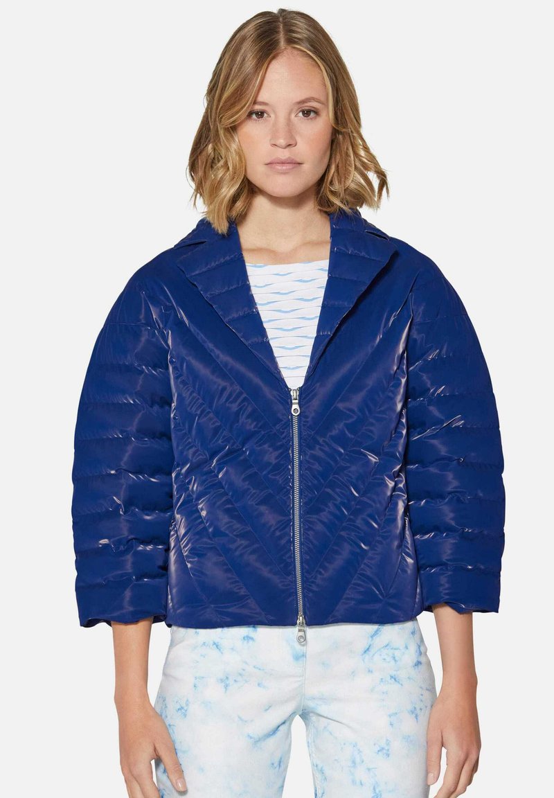 Marineblaue Steppjacke mit glänzender Oberfläche, gestepptem Design, Spitzkragen und Reißverschluss vorne. Kombiniert mit hellblauen gemusterten Hosen.
