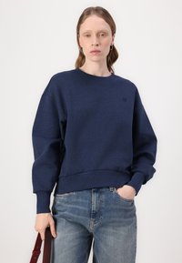 Mørkeblå genser med rund hals og ribbestrikkede mansjetter og kant, med en liten logo på brystet. Kombinert med lyseblå jeans.