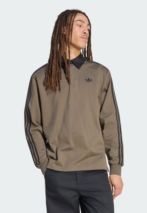 Homme avec de longues dreadlocks portant une chemise marron à manches longues avec un col noir et des rayures noires sur les manches, regardant sur le côté les mains dans les poches.