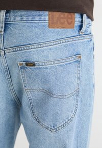 Light blue denim jeans with a brown leather label featuring "Lee." Includes a back pocket with a small black tag. Stitched details visible.