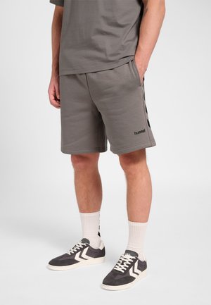 Mężczyzna w szarych casualowych szortach, pasującej koszulce, białych skarpetkach sportowych oraz szarych sneakersach z białymi wzorami w jodełkę stoi na białym tle.