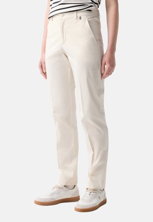 Personne portant un pantalon beige à jambes droites, des baskets blanches avec des semelles en gomme et une chemise rayée noir et blanc, debout sur un fond blanc.