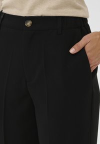 Un pantalon noir sur mesure présente un tissu lisse, un devant plissé, des poches latérales et une fermeture à bouton à la taille pour un ajustement sécurisé.