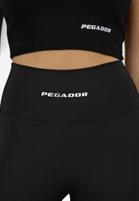 Melnā ribota krekla augšdaļa un augstu jostasviedru legingi, abi ar baltu "PEGADOR" zīmolu. Gluda tekstūra, pieguļoša forma, vienkrāsaini.