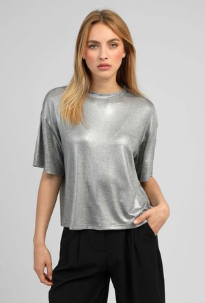 Jeune femme aux cheveux blonds et raides porte une chemise argentée brillante à manches courtes et un pantalon noir, debout devant un fond gris uni.