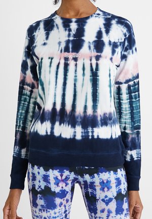 Osoba nosząca bluzę z długim rękawem w stylu tie-dye z granatowymi, białymi i różowymi wzorami oraz dopasowane niebiesko-białe spodnie z wzorem.