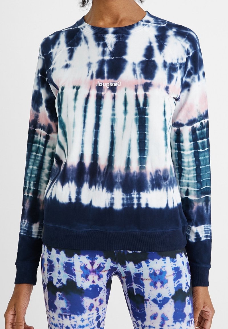 Personne portant un sweat à manches longues tie-dye avec des motifs bleu marine, blanc et rose, et un pantalon assorti à motifs bleus et blancs.