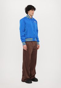 Chaqueta bomber de satén azul con puños y cuello a rayas amarillas, combinada con pantalones marrones de corte holgado y zapatos negros sin cordones.