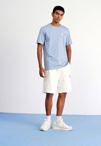 T-shirt en col rond bleu clair en coton, associée à un short couleur crème avec poches. Les chaussures sont des baskets de sport gris clair.