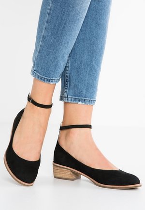 Czarne zamszowe baletki na niskim drewnianym obcasie z paskiem na kostce, z zaokrąglonym czubkiem i gładką teksturą, zestawione z krótkimi niebieskimi dżinsami.