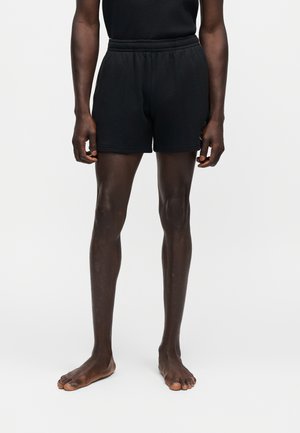 Blootsvoetse persoon staat, draagt zwarte getextureerde shorts en een bijpassende top, getoond van midden torso tot aan de voeten tegen een egale lichte achtergrond.
