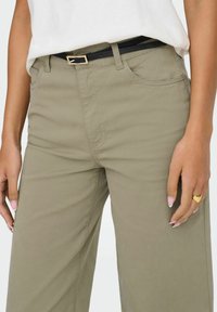 Femme portant un pantalon taille haute vert olive avec une ceinture noire, un haut blanc rentré et une bague en forme de cœur dorée à la main droite.