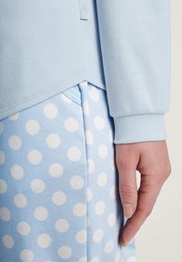 Lichtblauw langärmelig shirt met ribgebreide manchetten, gelaagd over lichtblauwe broek met stippen, toont soepele, zachte texturen en een afgeronde zoomdetail.