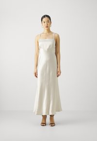 Satinová šaty na ramínka v off-white barvě, s tenkými ramínky a splývavým střihem. Černé sandály na podpatku doplňují outfit. Hladká textura.