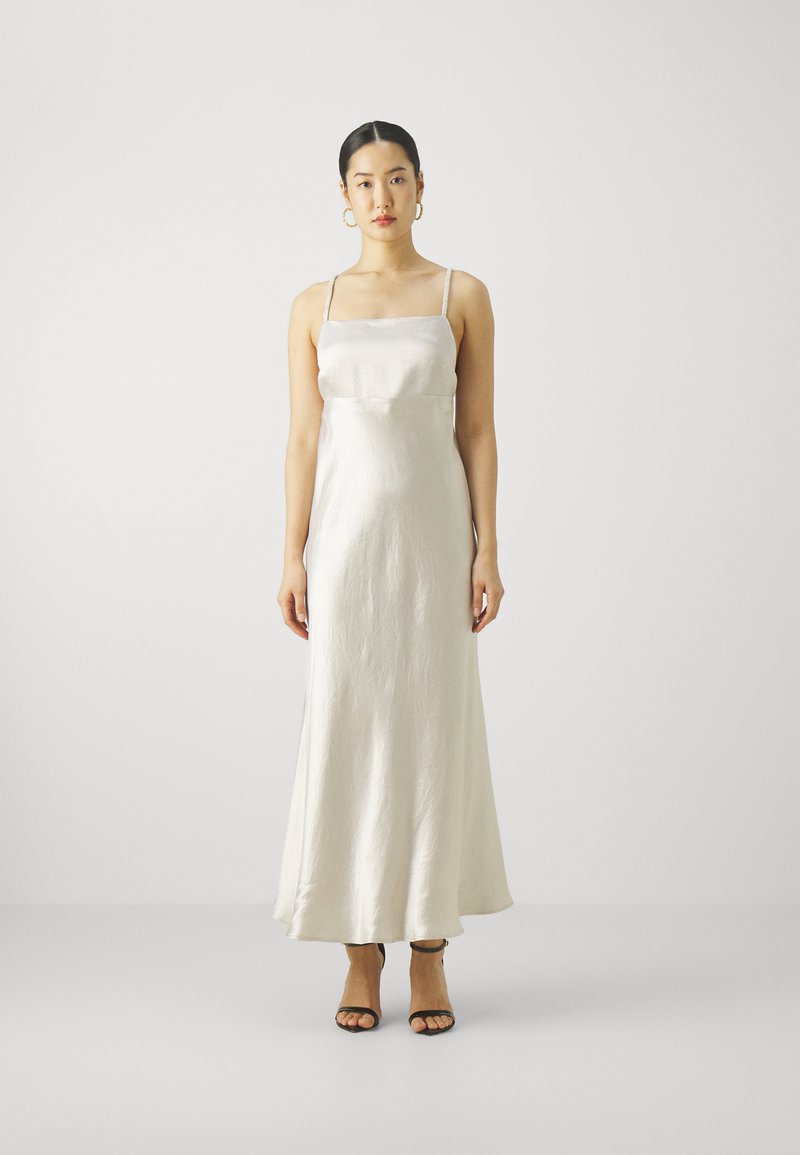 Satinová šaty na ramínka v off-white barvě, s tenkými ramínky a splývavým střihem. Černé sandály na podpatku doplňují outfit. Hladká textura.