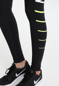 Leggings atletici neri con texture liscia, caratterizzati da strisce orizzontali verde fluorescente e un logo bianco lungo il lato. Indossate scarpe da ginnastica nere in rete.