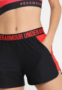 Pantalones cortos deportivos negros y rojos con tela texturizada, cinturilla elástica y bolsillos; cuentan con la inscripción "UNDER ARMOUR" destacada en rojo.