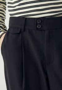 Melnas bikses ar klasikā piegriezuma, ar gludu priekšpusi, sānu kabatām un divām melnām pogām jostasvietā. Gluda auduma tekstūra.