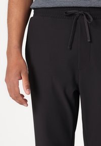 Sorte ribbede joggebukser med elastisk liv og snøring. Side lomme synlig. Modellens hånd hviler avslappet på siden.