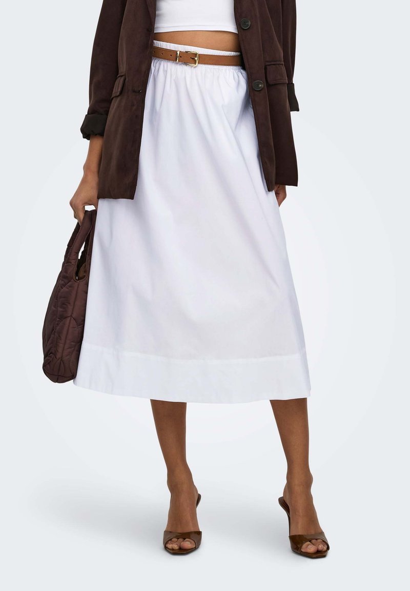 Femme portant une jupe midi blanche, un blazer court marron, une ceinture marron, des sandales à talons marron, et tenant un sac à main matelassé marron.