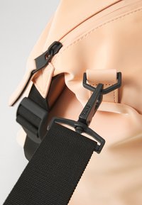 Stor stor ferskenfarget bag med svart maskinvare, med en teksturert stropp og slank design, som viser justerbar spenne og sømdetaljer.