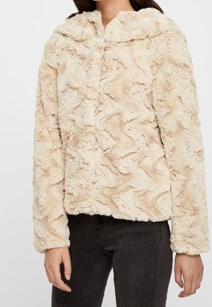 Femme portant une veste en fausse fourrure beige texturée à manches longues avec fermeture éclair devant, associée à un pantalon noir.