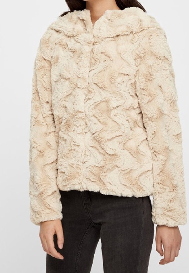 Femme portant une veste en fausse fourrure beige texturée à manches longues avec fermeture éclair devant, associée à un pantalon noir.