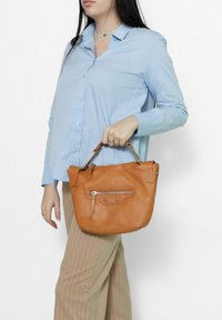 Borsa a mano in pelle color cuoio con forma arrotondata, dotata di una tasca con zip frontale e hardware in tonalità argento. Indossata con una camicia azzurro chiaro e pantaloni a fantasia.