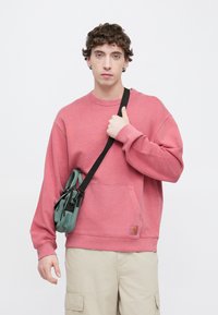 Ροζ φούτερ με μακριά μανίκια και τσέπη μπροστά, συνδυασμένο με πράσινη τσάντα χιαστί. Ομαλή υφή, καθημερινός σχεδιασμός.