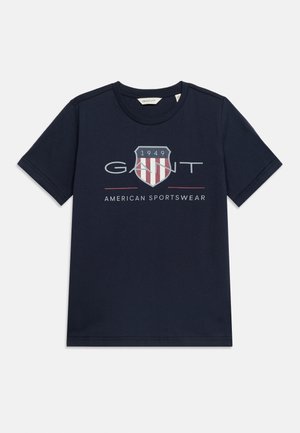 Μπλε ναυτικό κοντομάνικο t-shirt με το λογότυπο GANT, έμβλημα με ασπίδα και το 1949, καθώς και το κείμενο «American Sportswear» στο κέντρο του μπροστινού μέρους.