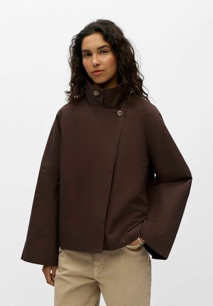 Femme aux cheveux bouclés portant une veste marron foncé à col montant et un pantalon beige sur fond blanc uni.