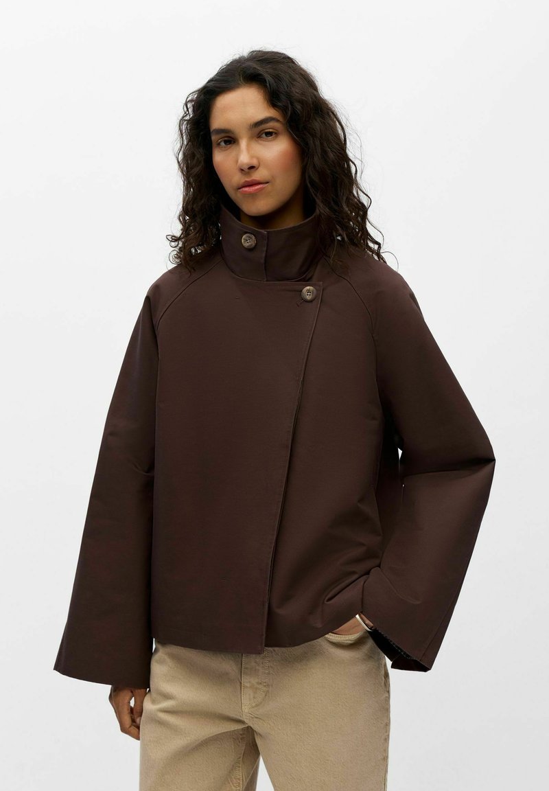 Femme aux cheveux bouclés portant une veste marron foncé à col montant et un pantalon beige sur fond blanc uni.