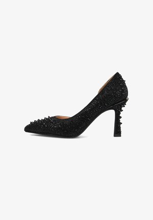 Zapato de tacón alto negro con punta afilada, adornado con brillantes remaches negros y un detalle texturizado en la superficie, con un tacón decorativo con picos.