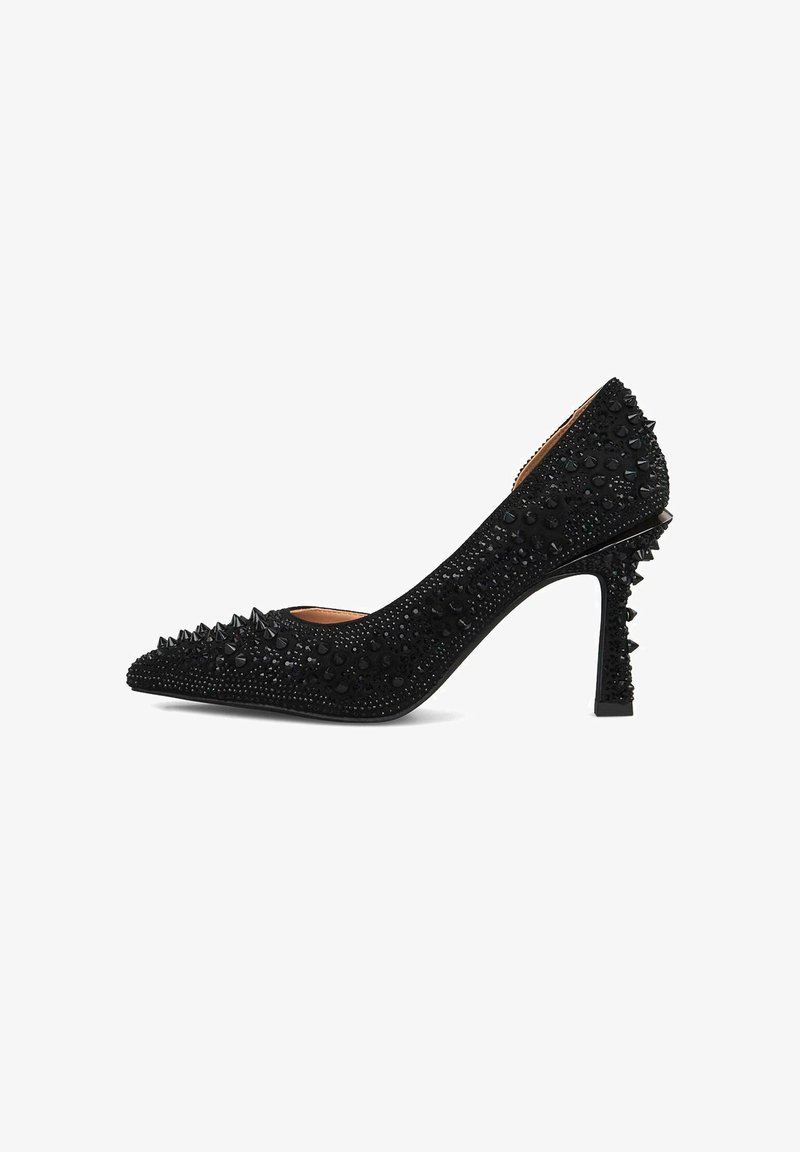 Zapato de tacón alto negro con punta afilada, adornado con brillantes remaches negros y un detalle texturizado en la superficie, con un tacón decorativo con picos.
