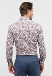 Camicia a maniche lunghe con bottoni di colore bianco con stampa paisley marrone; presenta un colletto classico e maniche con polsini. Abbinata a pantaloni blu.