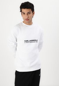 Bijela dukserica s crnim natpisom "KARL LAGERFELD 21 RUE ST-GUILLAUME PARIS", koja ima okrugli izrez i duge rukave.