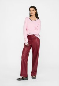 Rosa Strickpullover mit V-Ausschnitt und schwarzem Besatz, kombiniert mit hoch taillierten, weit geschnittenen bordeauxfarbenen Lederimitat-Hosen. Schwarze, verzierten flache Schuhe.