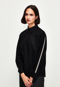 Camicia nera con bottoni e maniche lunghe, con una striscia argentata lungo il braccio sinistro. Realizzata in tessuto morbido con colletto classico.