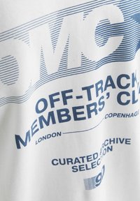 Baltas marškinėliai pagaminti iš medvilnės; ant jų yra mėlyna tekstas, sukurtas skirtingais šriftais ir linijų raštais, įskaitant "OFF-TRACK MEMBERS' CLUB" ir "CURATED ARCHIVE."