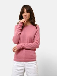 Růžový svetr z měkké, strukturované tkaniny s dlouhými rukávy, kulatým výstřihem a žebrovanými manžetami. Nosený s bílými kalhotami na neutrálním pozadí.