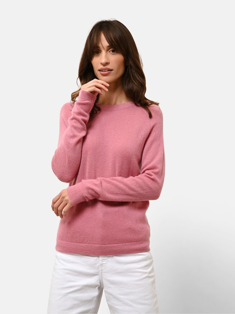 Růžový svetr z měkké, strukturované tkaniny s dlouhými rukávy, kulatým výstřihem a žebrovanými manžetami. Nosený s bílými kalhotami na neutrálním pozadí.