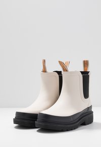 Botas Chelsea de goma beige con suelas negras, paneles laterales elásticos y tiradores con marcas en marrón y naranja. Superficie texturada, altura hasta la mitad de la pantorrilla.