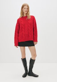 Maglione rosso lavorato a maglia con motivo a trecce, vestibilità oversize, maniche lunghe, abbinato a una gonna nera e stivali alti in pelle nera.
