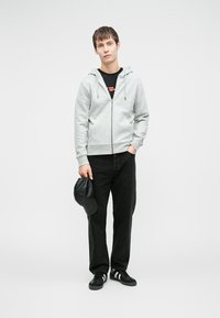 Šedá mikina na zip s bočními kapsami, černé tričko s červenými akcenty, černé džíny, černé tenisky a v ruce černá čepice.