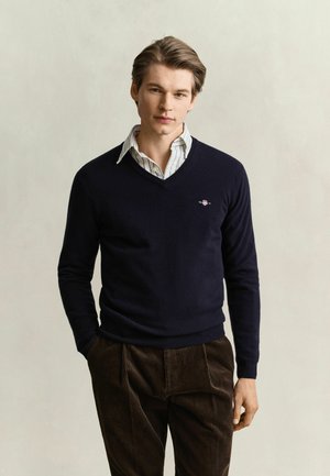 Jeune homme aux cheveux châtain clair portant un pull bleu marine à col en V par-dessus une chemise rayée et un pantalon en velours côtelé marron, debout devant un fond clair.