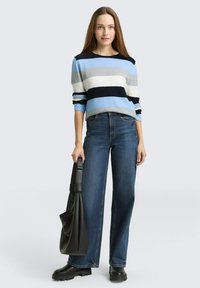 Blau-grau gestreifter Pullover mit weiten Ärmeln, kombiniert mit lockeren blauen Jeans. Das Modell hält eine schwarze Tasche und trägt klobige schwarze Schuhe.